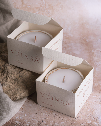 Die Verpackung der Veinsa Kerzen sind leicht geöffnet, so dass man die Kerzen sehen kann. Die eine Kerze steht auf einem Stein.